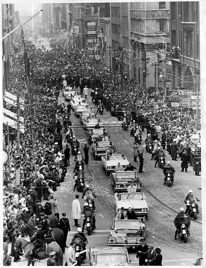 A black-and-white photograph of a crowded downtown street during a parade, confetti and streamers in the air, motorcycle escorts and open convertible cars moving through the crowd, overhead wires crossing the frame. Bay Street, Toronto.